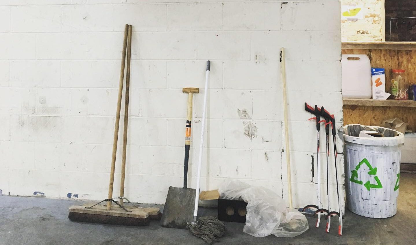 a broom, litter pickers, and shovel in front of a white wall in the Partisan basement