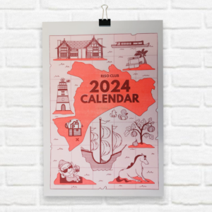 Partisan Calendar 2024 on white brick wall held up with a bulldog clip. The cover page features the silhuette of Salford with some landmark illustrations in spot colours wine red and fluorescent organge